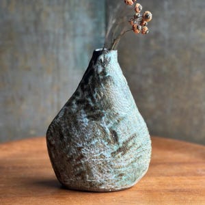 May include: A handcrafted ceramic vase with a unique, textured surface. The vase has a teardrop shape with a narrow neck and is adorned with a blend of blue, brown, and white hues. Dried, orange-toned berries add a natural touch.