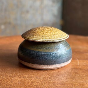 May include: A small, round ceramic lidded jar with a textured, speckled mustard-yellow lid. The base of the jar features horizontal bands of dark charcoal-gray and olive-green glaze, with a visible unglazed, light-colored clay rim at the bottom.