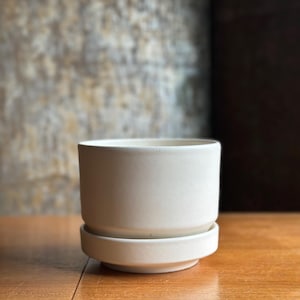May include: A white ceramic planter with a matching saucer. The cylindrical planter has a smooth, matte finish. The saucer is a low, circular base. The planter is designed for indoor or outdoor use.
