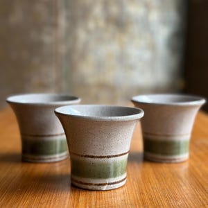 May include: Three ceramic vases with a flared top and a green band around the middle. The vases are beige with brown speckles and sit on a wooden surface. The vases are empty.