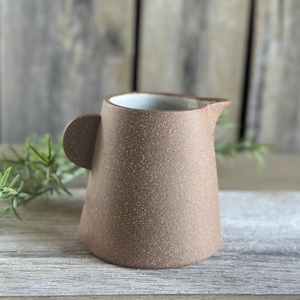 May include: A small, brown ceramic pitcher with a handle and spout. The pitcher has a textured surface and a white interior. It is sitting on a wooden surface with green foliage in the background.