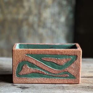 May include: A rectangular ceramic planter with a textured, terracotta-colored exterior. The planter has a green glaze design with abstract shapes.