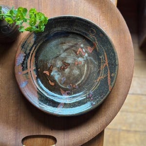 May include: A handmade ceramic plate with a dark, mottled glaze. The plate features a mix of brown, green, and blue hues with reddish-orange accents. The plate is round and appears to be approximately 8 inches in diameter, with a slightly raised rim.