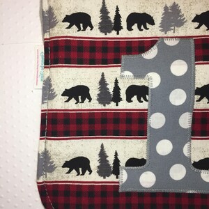 May include: A gray and white polka dot fabric bib with a red and black buffalo plaid pattern. The bib has a large gray fabric number one with white polka dots.