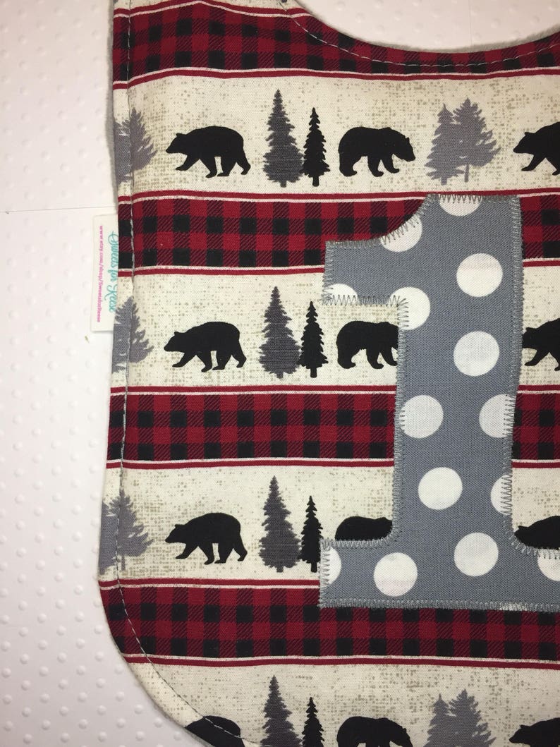 May include: A gray and white polka dot fabric bib with a red and black buffalo plaid pattern. The bib has a large gray fabric number one with white polka dots.