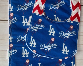 Puede incluir: Un conjunto de babero y babero para bebé en azul y blanco con un patrón de chevron rojo en el babero. La tela presenta un patrón repetido del logotipo de los Dodgers de Los Angeles con las palabras "LA Dodgers" y una pelota de béisbol.