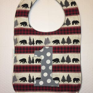 May include: A red and black plaid baby bib with a gray number one and white polka dots. The bib has a black bear and pine tree pattern.
