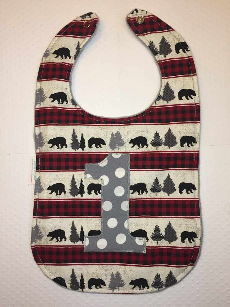 May include: A red and black plaid baby bib with a gray number one and white polka dots. The bib has a black bear and pine tree pattern.