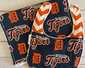 May include: A set of two baby items featuring the Detroit Tigers logo. A bib with a chevron pattern in orange and white and a burp cloth with the Detroit Tigers logo repeated on a navy blue background.