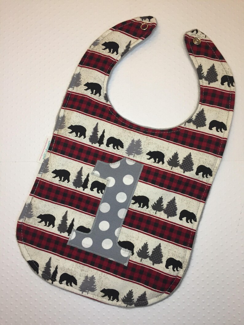 May include: A red and white plaid baby bib with a gray felt number one and a black bear print.