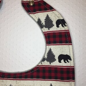 May include: A red and black plaid baby bib with a white background and a black bear and pine tree design. The bib has a soft, gray fleece lining and a snap closure.