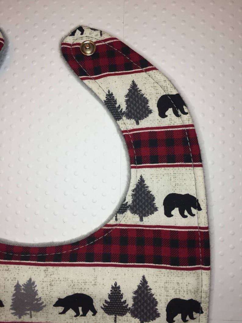 May include: A red and black plaid baby bib with a white background and a black bear and pine tree design. The bib has a soft, gray fleece lining and a snap closure.