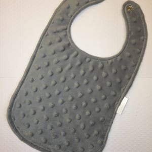 May include: A gray baby bib with a textured surface and two metal snaps. The bib has a white tag attached to the bottom right corner.