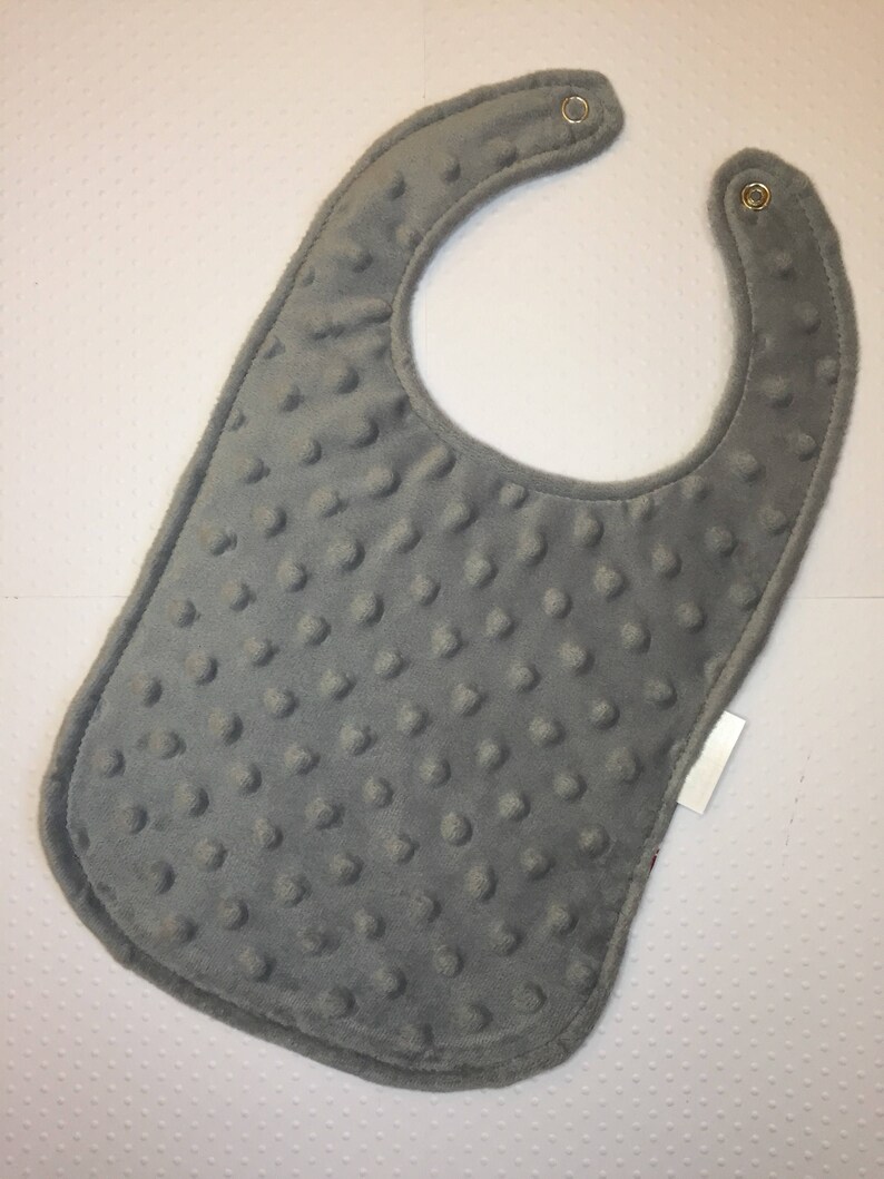 May include: A gray baby bib with a textured surface and two metal snaps. The bib has a white tag attached to the bottom right corner.