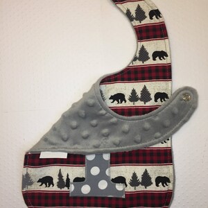 May include: A gray and red baby bib with a white and gray letter "I" and a woodland pattern of black bears and pine trees. The bib has a soft, gray fleece lining.