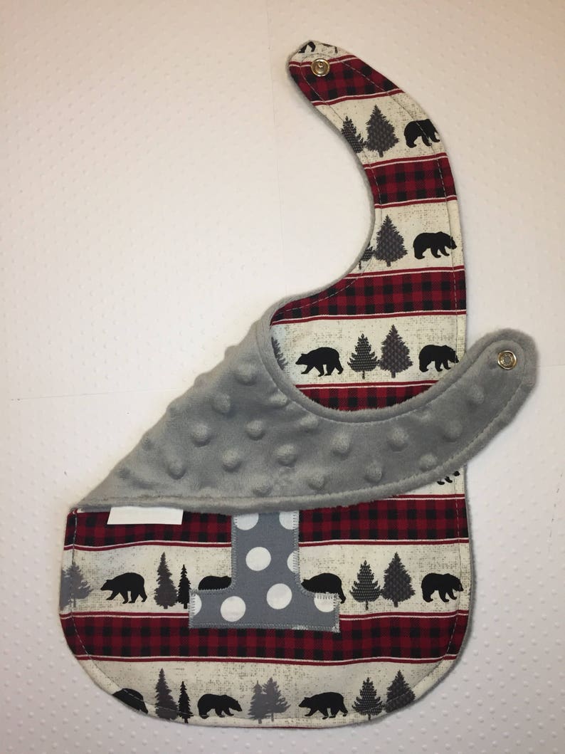 May include: A gray and red baby bib with a white and gray letter "I" and a woodland pattern of black bears and pine trees. The bib has a soft, gray fleece lining.
