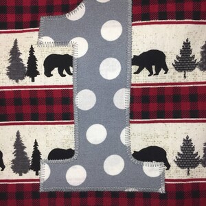 May include: A gray felt number one with white polka dots. The number is sewn onto a red and black plaid fabric with a white background.