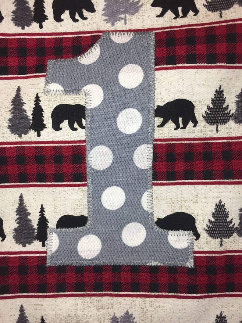 May include: A gray felt number one with white polka dots. The number is sewn onto a red and black plaid fabric with a white background.