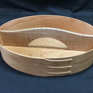 Pode incluir: Caixa oval de madeira estilo Shaker com acabamento castanho claro. A caixa tem um interior curvo e dividido e é construída com ripas de madeira e pequenos pinos espaçados. A caixa foi projetada para armazenamento.