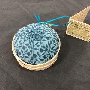 Pode incluir: Uma almofada redonda com um padrão floral azul, colocada numa base de madeira clara. Uma fita azul e um alfinete de segurança dourado estão presos. Uma etiqueta com texto também está presente. Um acessório de costura.