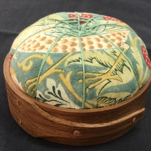 May include: A round pincushion with a wooden base. The cushion is covered in a patterned fabric with green, yellow, and red accents. Green thread secures the cushion. The wooden base has a curved design.