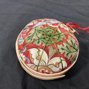 Pode incluir: Uma almofada redonda com um padrão floral em vermelho, verde e creme. A almofada é presa com fio verde e tem uma base de madeira. Uma fita vermelha e uma etiqueta estão presas.