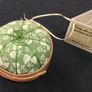 May include: A round pincushion with a light green floral fabric top, set in a wooden basket. The fabric is patterned with white leaves. A tag attached with a ribbon reads "Early Americana Workshop & Studio, New Hampshire."