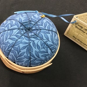 Pode incluir: Uma almofada redonda com tecido estampado floral azul claro, presa com linha azul escura. A almofada está em uma cesta de madeira clara. Uma etiqueta com o texto "Early Americana Workshop & Studio New Hampshire" está anexada.