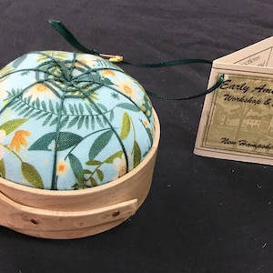 Pode incluir: Uma almofada redonda com um tecido floral azul claro, colocada numa base de madeira. O tecido apresenta flores brancas e amarelas com folhas verdes. Uma etiqueta diz "Early Americana Workshop & Studio, New Hampshire."