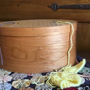 Pode incluir: Uma caixa oval de madeira castanha clara com tampa, com fio roxo claro e amarelo. A caixa está sobre um tecido estampado com desenhos florais. Um projeto de crochê está em andamento.