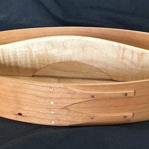 Pode incluir: Caixa oval de madeira estilo Shaker com acabamento castanho claro. A caixa tem um divisor curvo de madeira clara no interior. As laterais são unidas com pequenos pinos de metal decorativos. Um recipiente de armazenamento artesanal clássico.