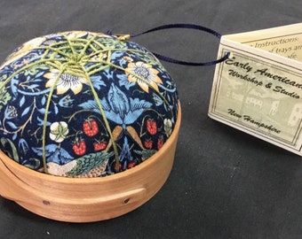 May include: A round pin cushion with a floral pattern in blue, yellow, and green, set in a light wood base. The cushion is secured with green thread. A tag reads "Early Americana Workshop & Studio, New Hampshire."