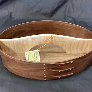 Pode incluir: Caixa oval de madeira estilo Shaker com um divisor interno de cor clara. A caixa é feita de madeira marrom escura com rebites de cobre e um divisor de madeira de cor clara. Uma etiqueta pende do divisor. A caixa é provavelmente usada para armazenamento.