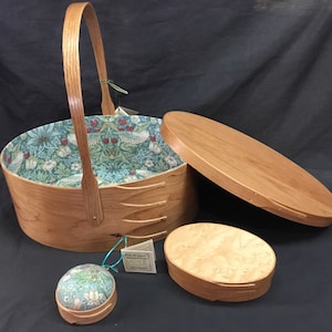 May include: A wooden sewing basket with a handle, open lid, and a patterned fabric interior. A small wooden box and a pin cushion are also present. The basket and boxes are made of light-colored wood.