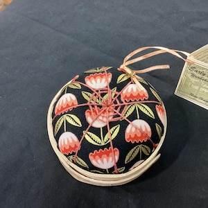 Pode incluir: Uma almofada redonda com um padrão floral em vermelho, branco e dourado sobre um fundo preto. A almofada está em uma base de madeira clara. Uma etiqueta com texto é presa com uma fita rosa.