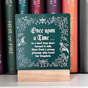 May include: A green chalkboard sign with white lettering that reads "Once upon a Time... in a land long since burned to ash, there lived a young princess who loved her kingdom." The sign is sitting on a wooden stand in front of a bookshelf.