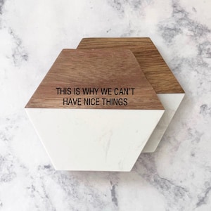 May include: Two hexagonal coasters with a white marble top and a brown wood bottom. The top coaster has the text "THIS IS WHY WE CAN'T HAVE NICE THINGS" printed in black.