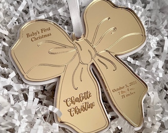 May include: Gold-colored mirrored ornament shaped like a bow with the text "Baby's First Christmas" and "Charlotte Christine" along with the date "October 5, 2023", weight "7 lbs. 4 oz." and length "21 inches".
