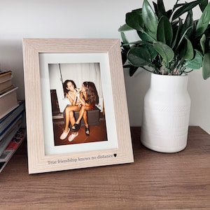 Op de afbeelding: Een licht houten fotolijst met een foto van twee vrouwen die naar elkaar lachen. De lijst heeft de tekst "True friendship knows no distance" en een klein hartje.