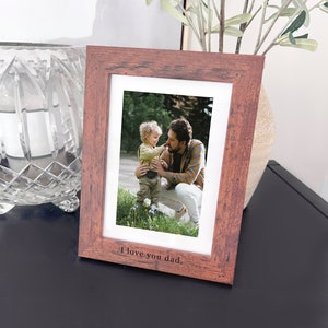 May include: A rustic wooden picture frame with a photo of a father and son. The photo shows the father looking down at his son, who is smiling. The frame has the words "I love you dad." engraved on the bottom.