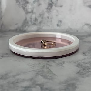 May include: A small, round, white and pink tray with a rose gold interior. A gold ring with a black stone is sitting on the tray.