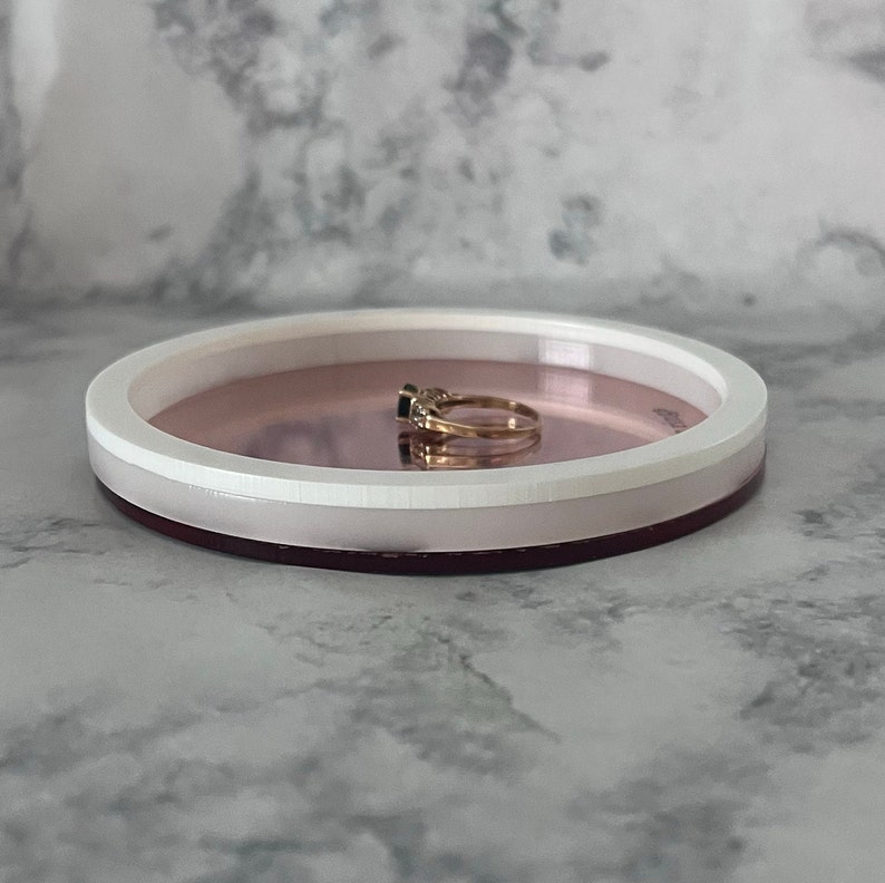 May include: A small, round, white and pink tray with a rose gold interior. A gold ring with a black stone is sitting on the tray.