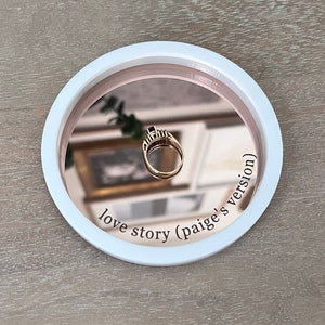 May include: A white and rose gold ring dish with a mirrored surface. The dish has the text "love story (paige's version)" inscribed on the rim.