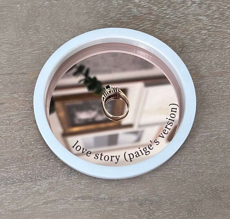May include: A white and rose gold ring dish with a mirrored surface. The dish has the text "love story (paige's version)" inscribed on the rim.