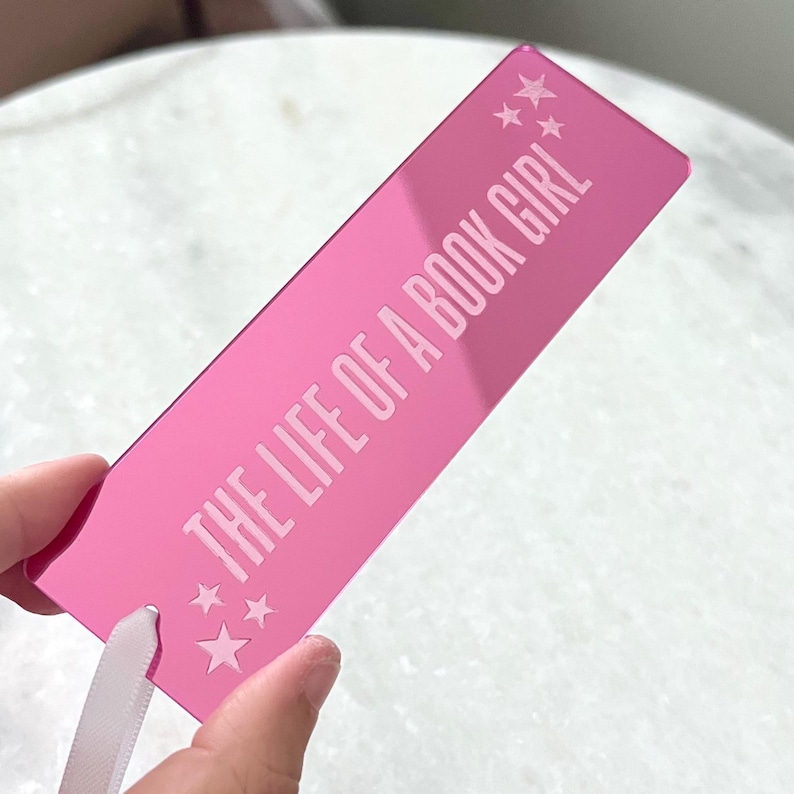 May include: A pink bookmark with the text "THE LIFE OF A BOOK GIRL" in white. The bookmark has a ribbon attached and star graphics. The bookmark is rectangular and appears to be made of a reflective material.