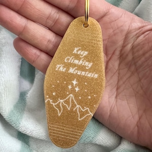 May include: Gold-colored keychain shaped like a hotel key with the text "Keep Climbing The Mountain" and a white outline of mountains and stars.