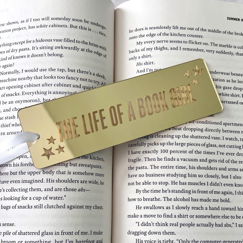 May include: Gold-colored metal bookmark with the text "THE LIFE OF A BOOK GIRL" and three star designs. The bookmark is rectangular with a ribbon attached, resting on the open pages of a book.