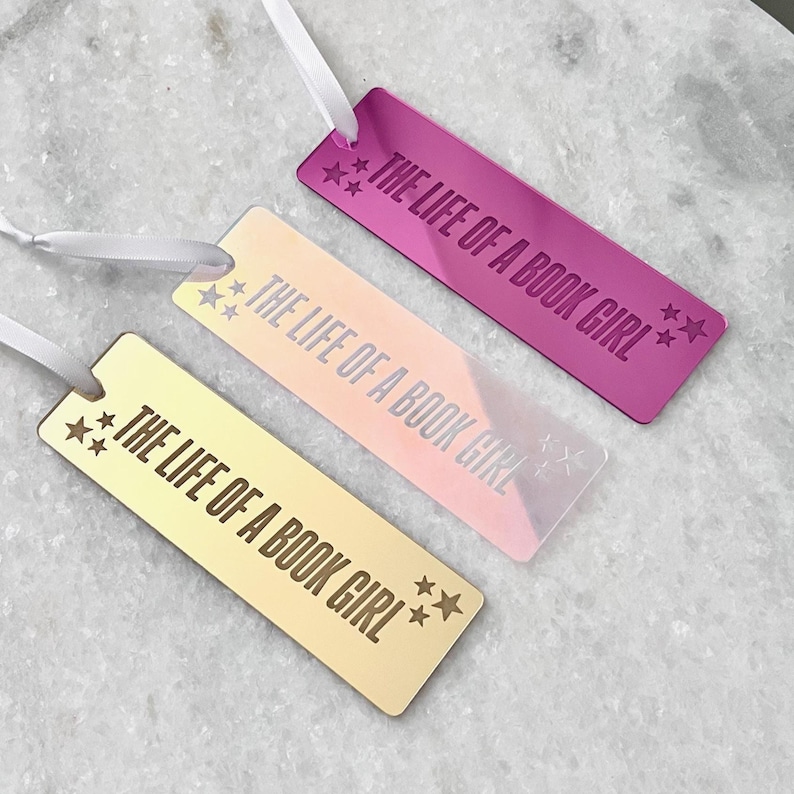May include: Three metallic bookmarks in pink, gold, and peach, each with the text "THE LIFE OF A BOOK GIRL" and star accents. White ribbon is attached to each bookmark. The bookmarks are displayed on a marble surface.