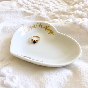 May include: White heart-shaped ceramic ring dish with gold lettering that reads "forever & always". A gold ring with a black stone is sitting in the dish.