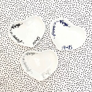 May include: Three white heart-shaped ceramic dishes with black and silver lettering. The dishes are decorated with the words "always & forever" and "always & married" and the initials "A-B", "M-B", and "C-F".